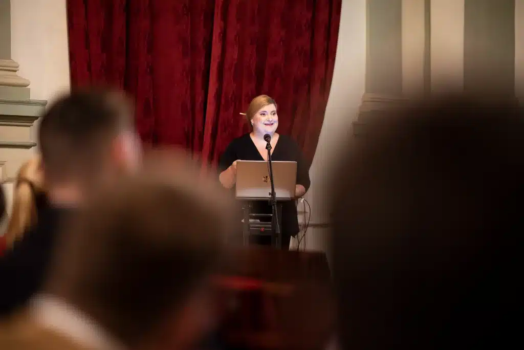 Female presenter giving a presentation to colleagues seated at a table with notes and laptops.