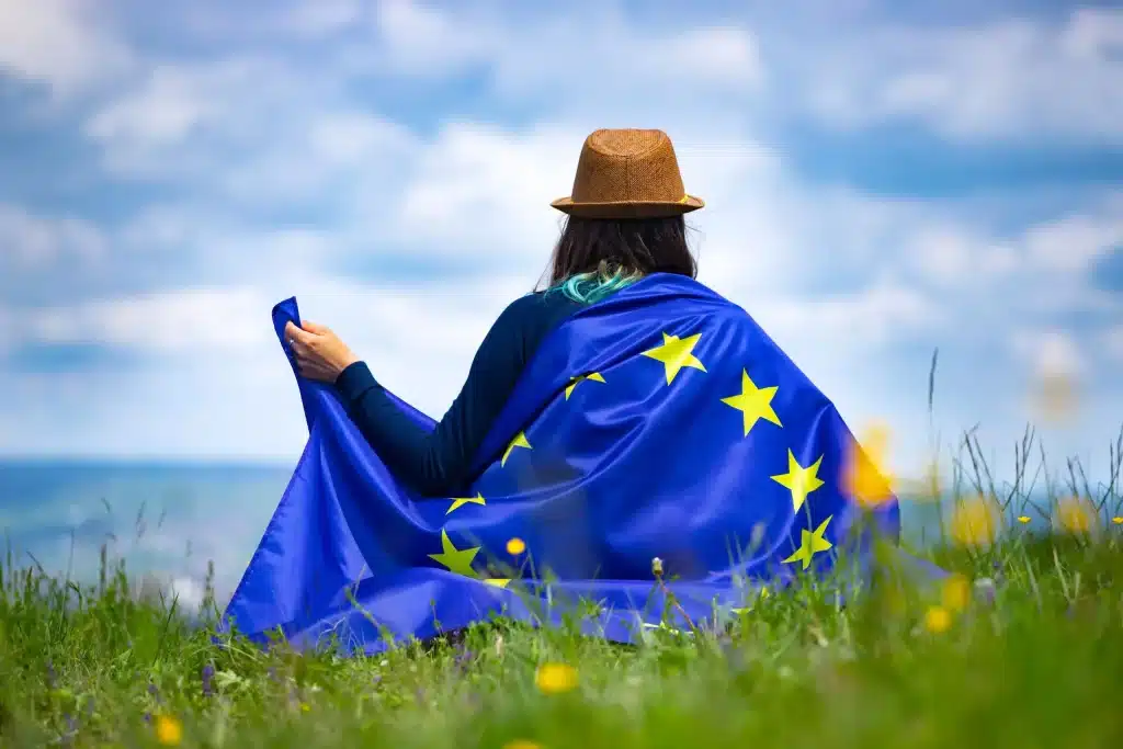 Háttal ülő személy kalapban, vállán az Európai Unió zászlajával a mezőn.