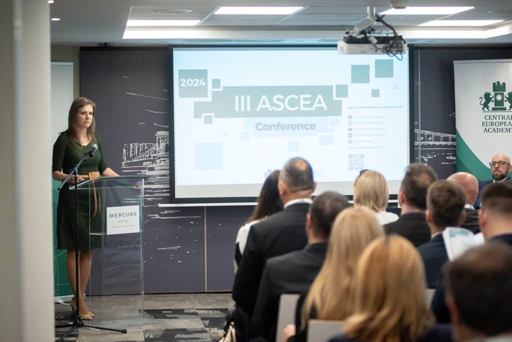 Nő a színpadon előadást tart, a háttérben a III. ASCEA konferencia vetítése látható.