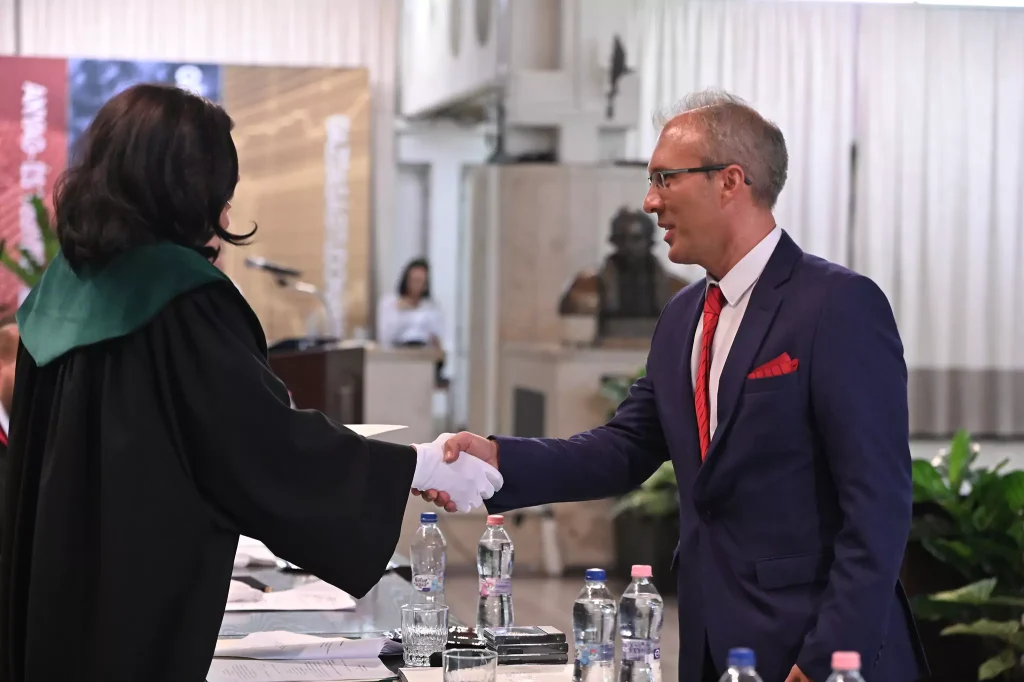 Ünnepélyes díjátadón egyetemi professzor kezet fog egy elegáns öltönyös férfival.
