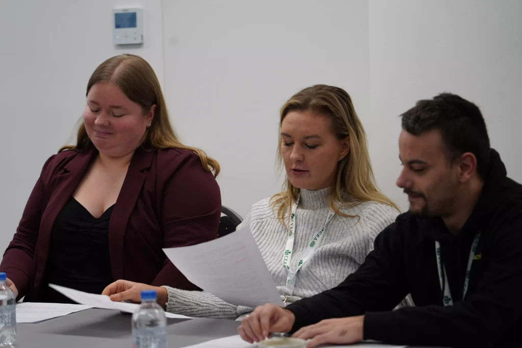 Two women and a man sit at a table reading documents and taking notes, with bottled water in front of them.