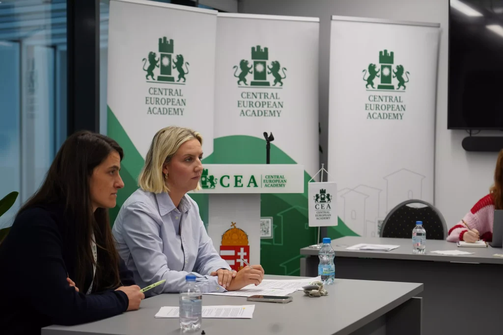 Two women sit in the conference room in front of Central European Academy banners, with notes and bottled water on the table.