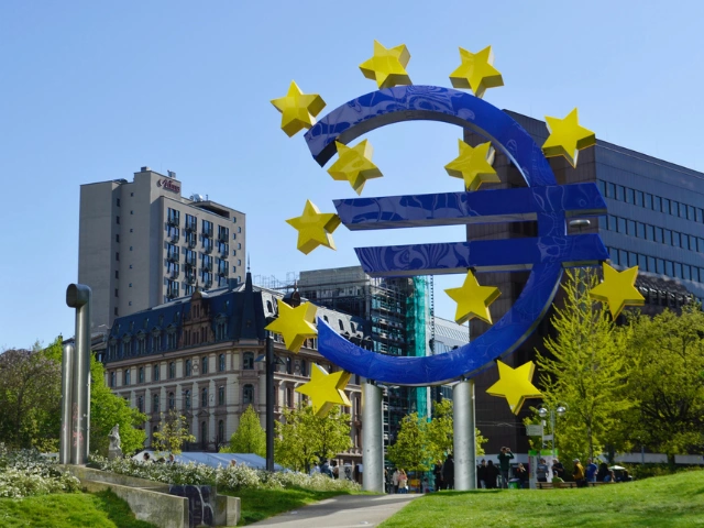 Az Európai Központi Bank ikonikus kék euró jel szobra arany csillagokkal, frankfurti városi háttérrel.
