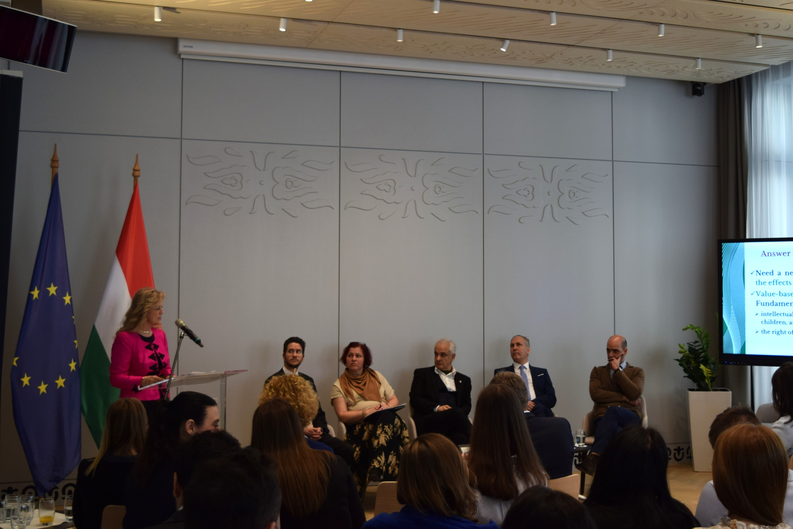 Speaker at a podium with five seated panel members and a screen, with the EU and Hungarian flags in the background.